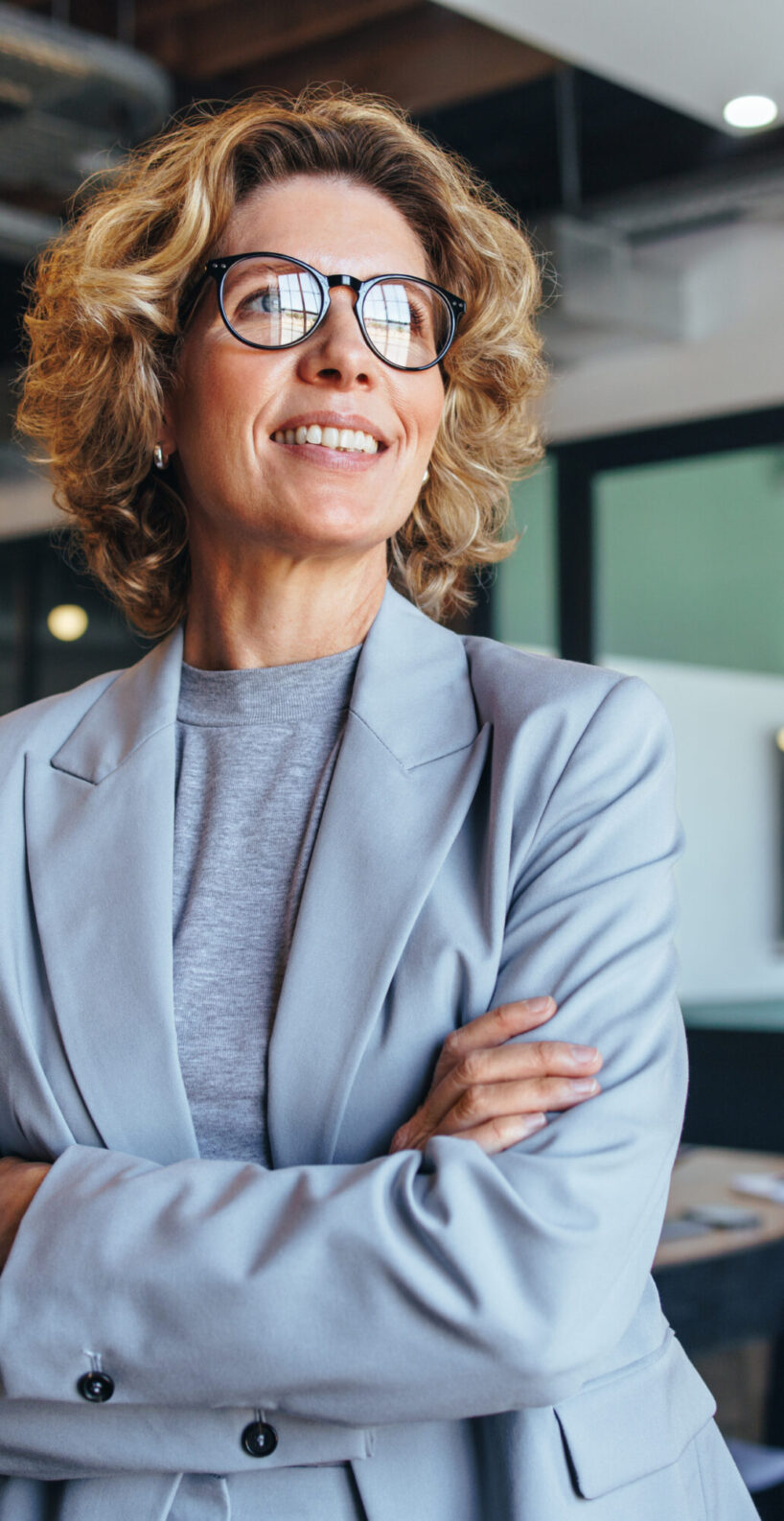 Business woman in her 40's looks away thoughtfully in an office. Professional woman standing in a suit with crossed arms.