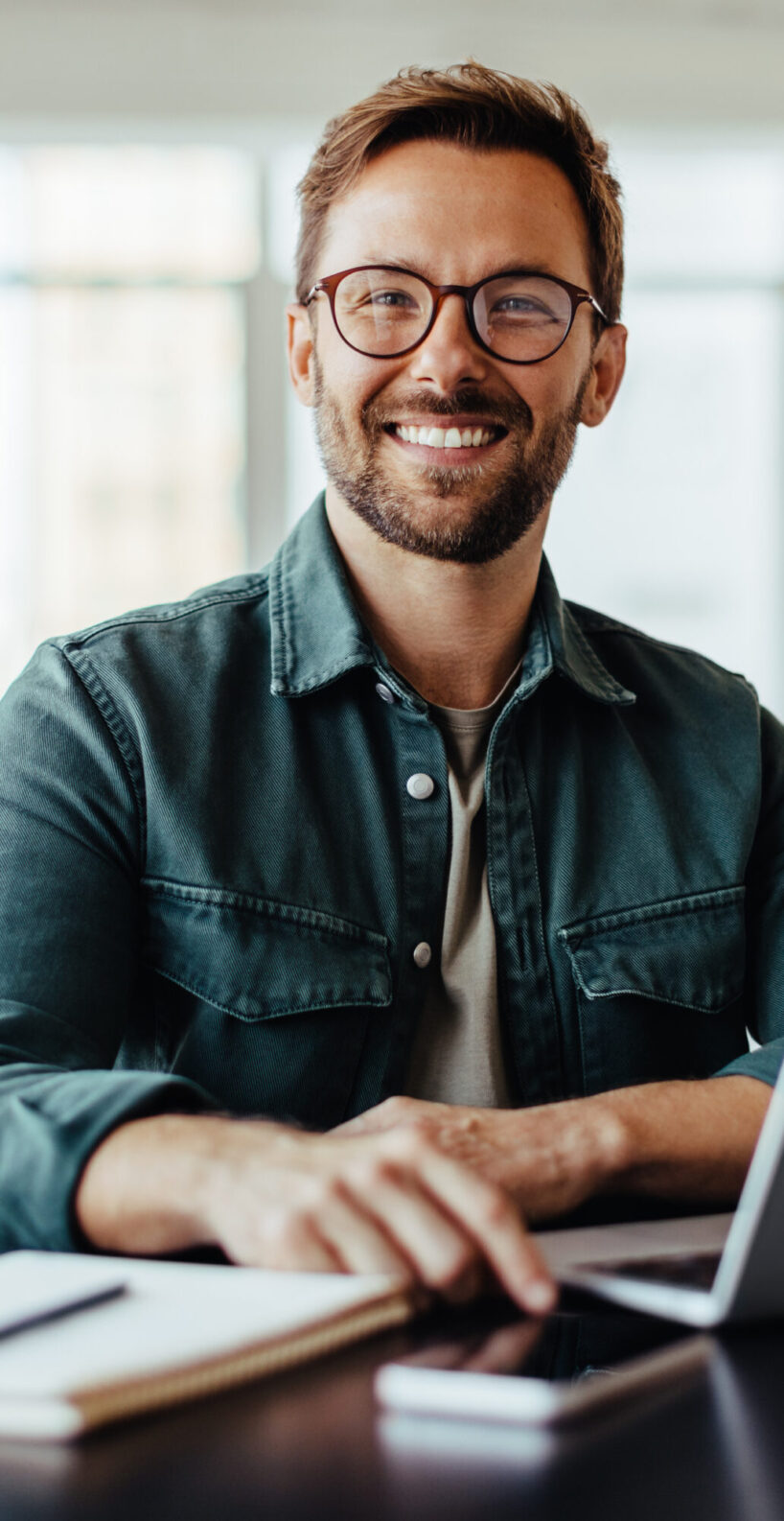 Portrait of a business man sitting in an office with his colleagues in the background. Happy business man working in a co-working office.